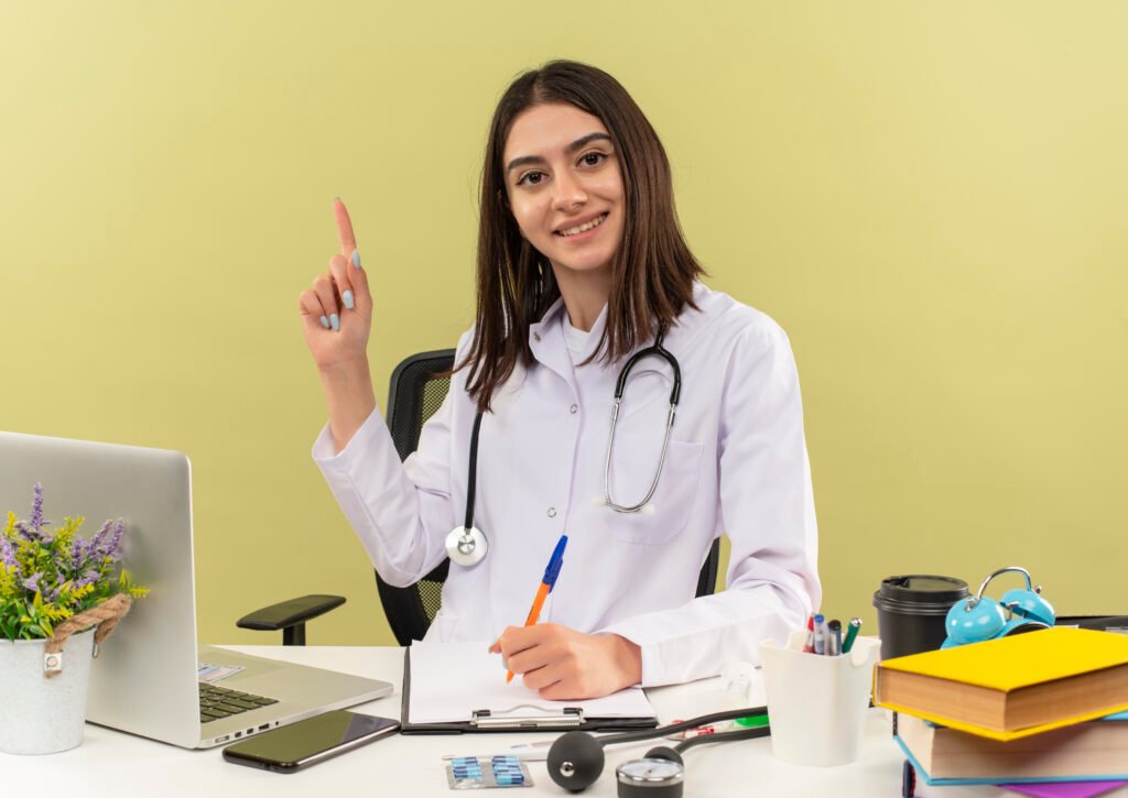 young woman doctor in white coat with stethoscope around her neck showing index finger having new idea sitting at the table with laptop over light background